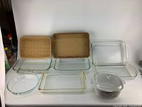Overview of 9 kitchenware items: clear glass Pyrex baking dishes in various sizes and 2 woven serving trays, all with vintage signs of use and wear.