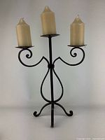 Full view of vintage triple candlestick holder with 3 ivory candles, showing hand forged iron frame and decorative scrolls.