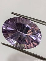 Close-up photo of an oval cut genuine amethyst gemstone held by tweezers, showing its rich purple color and faceted surfaces.