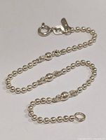Silver beaded bracelet arranged in a looping pattern against a white background showing the clasp and tag details.