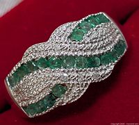 Close up image of silver ring showing rows of round emerald stones and clear cubic zirconia stones in a twisted pattern on the band.