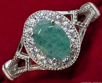 Close-up front view of silver ring featuring an oval green emerald center stone with surrounding clear cubic zirconia stones in a halo setting, on a red background.