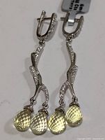 Pair of silver drop earrings featuring two light green carved green amethyst teardrop gemstones each suspended from a silver stem embellished with small clear stones.