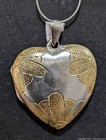 Close-up of front side of the silver heart-shaped locket showing embossed gold-tone leaf patterns and bail attached to silver chain.
