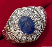 Front angled view of silver ring showing oval blue sapphire center surrounded by clear cubic zirconia stones and engraved detailing on band.