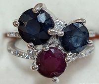 Close-up front view of silver ring with two blue sapphire stones, one red ruby stone, showing faceting and prong settings.