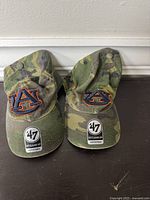 Two camouflage style baseball caps with embroidered Auburn University AU logos on front, viewed from above showing logos and original branded stickers.