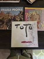 Photo showing three vinyl records laying flat on black surface: Village People Cruisin with iconic desert/construction cover, M Pop Muzik with baby on pink cover, and Toto album with plain white heavily worn cover with black/red markings.