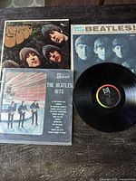 Three Beatles vinyl albums pictured including a sealed Plastic wrapped Spanish Odeon pressing called The Beatles Hits, and exposed U.S. Capitol Rubber Soul and Meet the Beatles! albums.