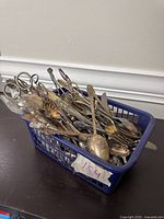 Photo of assorted vintage silverware in blue plastic basket showing spoons, forks, and other utensils with tarnish and wear.