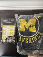 Closed tin case with set of 28 double-six dominoes showing maize M logo and sealed navy blue Michigan Wolverines T-shirt with large M and 'Wolverines' text.