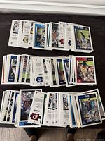 Photo showing multiple stacks of NFL trading cards laid out flat on a dark surface with front and back sides visible, showing various players from different teams in vibrant colors and clean condition.