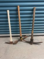 Three heavy-duty pickaxes with wooden handles and dual-point metal heads leaning on a metal background. One handle is painted white with a crack; others are natural wood with wear.