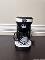 Front view of the Star Wars single-serve coffee maker with mug placed on the drip tray showing the Rebel Alliance logo and Star Wars logo on the machine.