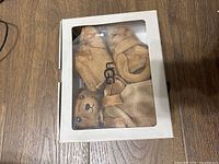 UGG gift box containing the baby booties and plush bear, view through clear window shows brown booties and bear inside.