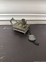 Front and side view of the vintage miniature metal oil lamp showing detailed floral and decorative patterns, handle, and hanging emblem tag.