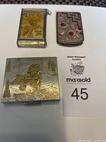 Lot showing three vintage compact cases on white surface: leather floral, New York metal map, and shell patterned compact.