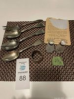 Three silver plated spoons, one meat fork, a sterling silver napkin ring, a silver plate cleaner, and salt and pepper shakers with mother of pearl tops and cut glass bodies arranged on a textured mat with packaging.