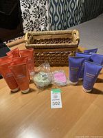 Six orange tubes of firming body cream and five purple anti-cellulite gel tubes arranged in a semi-circle around a woven basket; also visible are silicone scalp massager and nail buffer in packaging on table