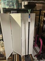 Full view of the black floor lamp showing its height and bowl-shaped shade, placed next to a white folding screen and pink exercise equipment.