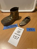 Photo showing both brass shoe-shaped ashtrays side by side on a surface with size indication tape and auction tag.