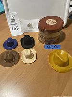 Overall lot with Stetson branded tin and five male doll hats of 1/6 scale in various colors and styles on wooden table.