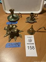 All four items in the lot: three brass candlesticks and one candle snuffer shown from above and side angles on wooden surface with auction tag and measurement guides.
