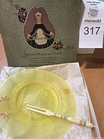 Lemon plate and fork displayed in original box with protective tissue paper, showing plate's translucent yellow color and fork with white handle and colorful bands.