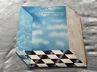 Front cover of 'The Low Spark of High Heeled Boys' album by Traffic showing sky and cloud-themed artwork with checkerboard pattern border; cover shows signs of wear and edge damage.