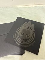 Front of black square card showing embossed grey cat face with cityscape and Meow Meow Industries text.