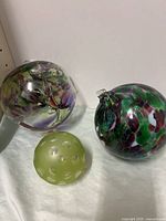 Three decorative glass balls on white surface: two large, colorful blown glass balls and one small matte green cast glass ball with raised bumps and hanging loop.