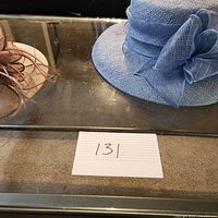 Both hats displayed, beige on left with brown loops and light blue on right with large fabric bow, on glass surface.