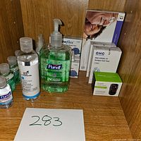 Photo showing several bottles of hand sanitizer including Purell and anafect brands, Flowflex and OHC COVID-19 Antigen Self Test kits in boxes, plus a glucose meter box on a wooden shelf.