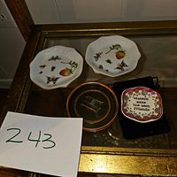Top view of the set showing two white porcelain small plates decorated with strawberries, butterflies, and insects, one bronze/brass coaster holder with engraved chair design, and one square ceramic plate with floral border and inscription, all displayed on a glass surface.