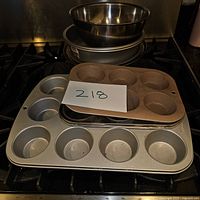 Photo showing set of baking pans stacked, includes two muffin pans (6 and 12-cup), three metal pie/cake pans, and two metal mixing bowls on top.
