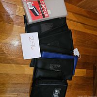 Photo showing multiple soft-sided banker bags in black and blue, some with clear ID windows, arranged on a wooden floor alongside a new Staples poly expanding check file in packaging.