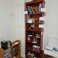 Front view of tall wooden bookshelf filled with books and other items (contents are not included in sale). Seven shelves visible with various objects labeled with paper tags.