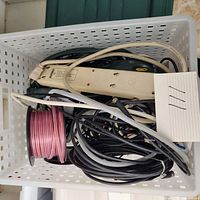 Top-down view of box showing assorted extension cords, power strips, and speaker wire spool in a white plastic basket.
