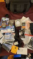 Wide shot showing black camera carrying bag, various camera accessories scattered including lens cap buckles, cables and remote shutter, with handwritten lot number card.