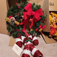 Photo of the Christmas wreath with red bow, berries, pine cones, and gold presents visible on a carpeted floor with box background.