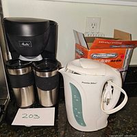 Melitta coffee maker with two stainless steel travel mugs and white Proctor Silex electric kettle on kitchen counter.