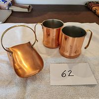 Photo showing two copper mugs and a small copper basket with handle placed on a towel with lot number 62.