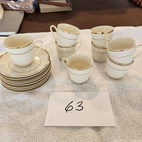 Full set of 12 white porcelain demitasse espresso cups and 12 matching saucers arranged on a towel. Cups and saucers decorated with fine gold trim on rims and edges.