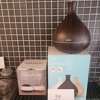 Brown wood grain Anjou aroma diffuser placed on the box of the new diffuser. The new diffuser is shown in the box with images depicting white and wood finishes.