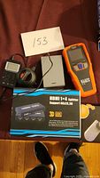 Photo of electronics lot showing HDMI splitter box, tape measure, network cable tester, power bank, handheld digital device, and security camera box.