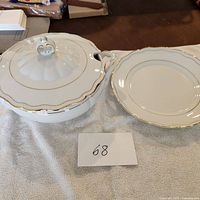 White porcelain soup tureen with gold trim next to matching oval platter, both on white cloth, showing detail and condition