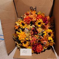 Fall wreath inside a cardboard box showcasing mixed artificial flowers and leaves in autumn colors.
