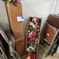 Box with assorted faux flower stems in red, white and cream colors, some leaning outside the box, on concrete floor near stacked folding chairs and tables.