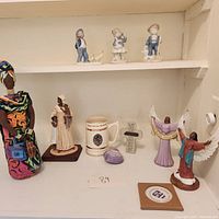 Overview of various figurines and collectibles arranged on a shelf and surface: porcelain children figurines, angel figurines, African woman figurine, Southern belle figurine, beer stein, shell, cross plaque.