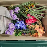 Wide shot of bin containing assorted artificial flowers including orange sunflowers, pink roses, purple bell flowers, blue flowers, and green foliage stems.